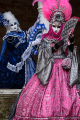 Fototapeta na wymiar Masked women in ornate blue and pink costumes standing next to stone column in a courtyard during the carnival (Carnival di Venezia)