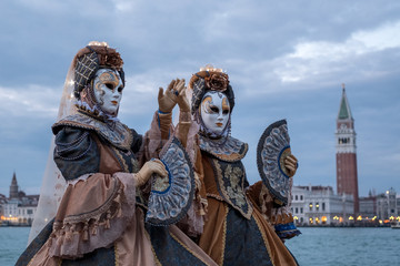 Obraz premium Venice Italy, February 2018. Two women in traditional costumes and masks, with decorated fans, standing in front of the Grand Canal with San Marco in the background, during the Venice Carnival