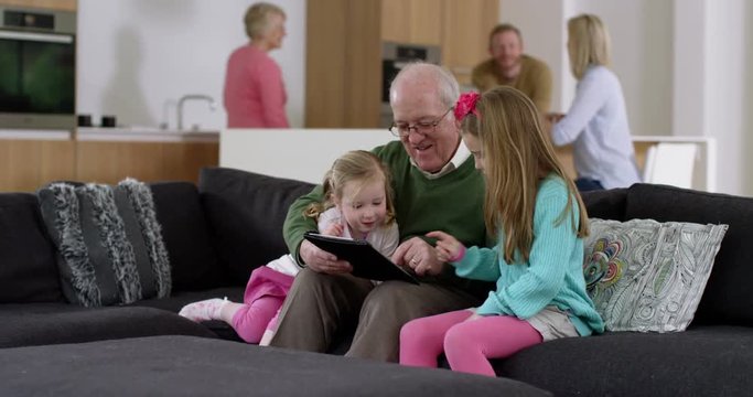 Grandfather plays game on electronic tablet with grand daughters in beautiful house whilst parents chat in background