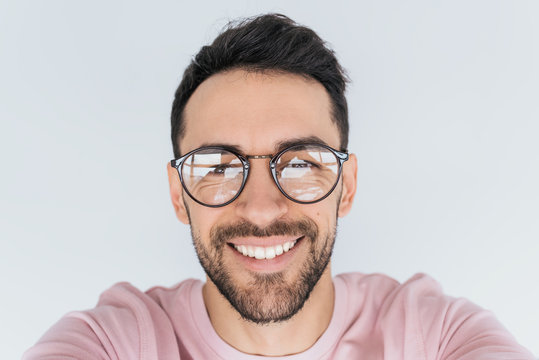 Closeup portrait of handsome young happy smiling bearded male model look at the camera, wears round trendy glasses with toothy smile. Cheerful man in spectacles, pink clothes advertises in studio wall