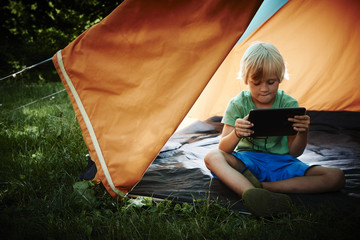 Cute child boy playing on tablet at sunset in campside, in the tent

