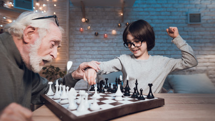 Grandfather and grandson are playing chess together at night at home. Boy is winning.