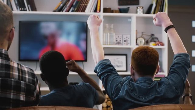 Multi-ethnic men celebrate goal together. Back view. Excited fans watch football game on the couch together. Emotion.