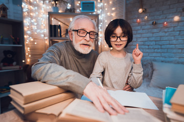 Grandfather and grandson are doing homework at night at home. Granddad is helping boy.