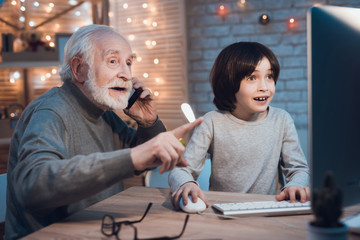 Grandfather and grandson are playing games on computer at night at home. Granddad is cheering for boy.