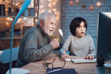 Grandfather and grandson are playing games on computer at night at home. Granddad is cheering for boy.