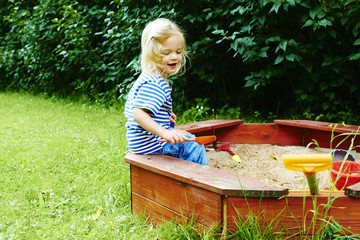 Happy little child, adorable blond child  girl having fun playing outdoors in the garden with plastic toys sitting in blue sand box on a sunny summer day © Petr Bonek