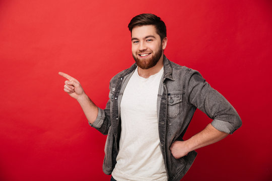 Photo of handsome cheerful man 30s in jeans jacket gesturing finger aside on copyspace with gladness on face, isolated over red background