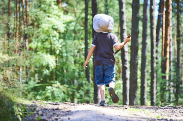 Small child boy walking or hiking on a path through the forest in summer with sun hat on head
