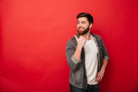 Photo of handsome man 30s in casual clothing gesturing fingers backward on copyspace with happy smile, isolated over red background