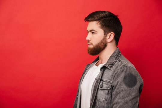 Portrait in profile of concentrated man 30s in jeans jacket looking straight on copyspace with brooding serious gaze, isolated over red background