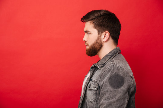 Portrait in profile of concentrated bearded man in jeans jacket looking straight on copyspace with brooding serious gaze, isolated over red background
