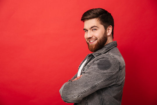 Half turn portrait of attractive man in jeans jacket posing on camera with amazing smile and arms folded, isolated over red background