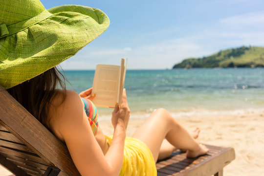 Side view of a young beautiful woman reading a book while sitting on a wooden lounge chair at the beach in a sunny day during summer vacation in Flores Island, Indonesia