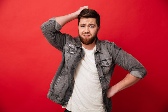 Upset man 30s in jeans jacket looking on camera and scratching his head in perplexity, isolated over red background