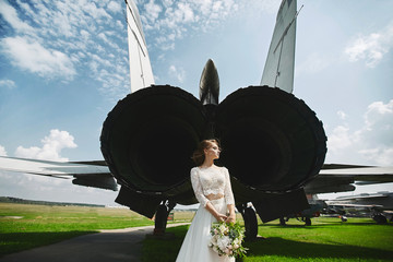 A young beautiful brown-haired bride is standing behind a jet plane