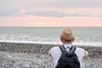 Young man in hat and with backpack sitting on the beach. Sea and sunset sky. Back view