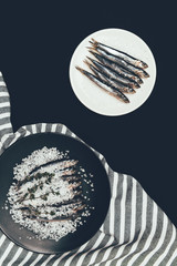Salted pile of salted fish with rosemary in frying pan and plate with fish isolated on black