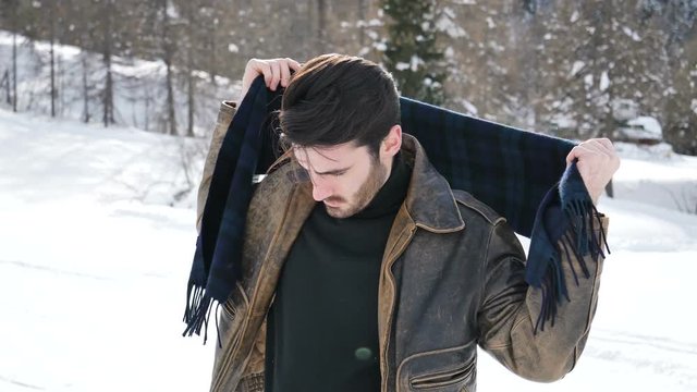 Stylish man putting on wool scarf and confidently looking away on territory of contemporary winter resort covered in snow in the mountain
