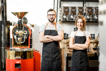 Portrait of a couple of baristas in uniform standing together in the coffee shop with coffee roasting machine on the background