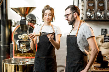 Couple of baristas in uniform checking the quality of roasted coffee beans standing near the roaster machine