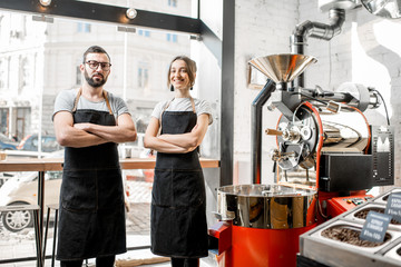 Portrait of a couple of baristas in uniform standing together in the coffee shop with coffee roasting machine on the background