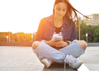 A beautiful girl in a jeans jacket, sitting on the steps on a city street, uses her smartphone. A woman communicates through the Internet. Lifestyle. Trend. Hipster.