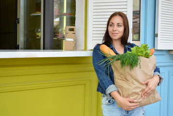 A smiling girl in a jeans jacket is holding a paper bag with food in her hands. The woman stinted in the supermarket. Crafting. Baguettes. Leaves of lettuce. Carrot.