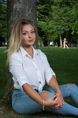 Attractive young girl sits in a park near a tree. Blond girl in free city clothes. White shirt. Sexy.