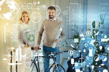 Important choice. Cheerful young engineer holding a rubbish bin with a recycling sign on it while her positive colleague putting his hand on the bike and smiling