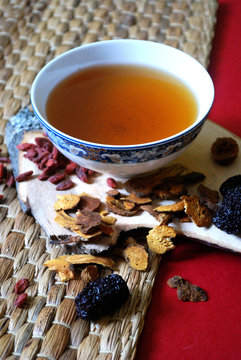 Goji berries, chinese dates, astragalus root pieces with a bowl of herb tea on red background. Vertical photo.