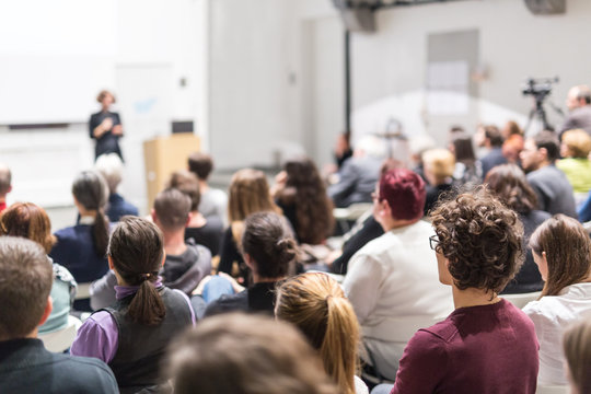 Female Speaker Giving Presentation In Lecture Hall At University Workshop. Audience In Conference Hall. Rear View Of Unrecognized Participant In Audience. Scientific Conference Event.