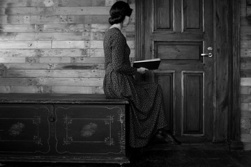 Beautiful woman sits in profile on a black chest and reads a book on a wooden background