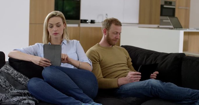 Attractive couple have uncomfortable glance at each other after argument, whilst looking at tablets in modern contemporary home,