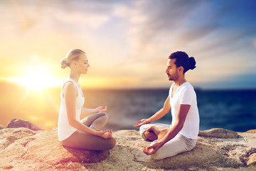 Fototapeta na wymiar yoga , mindfulness, harmony and people concept - happy couple meditating in lotus pose outdoors over sea background