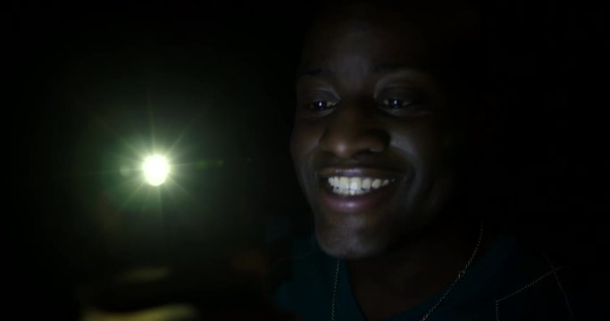 BCU dark skinned man talking on video call through cellphone at night, looks from screen to viewer and smiles. Hand-held 4K with lens flares.
