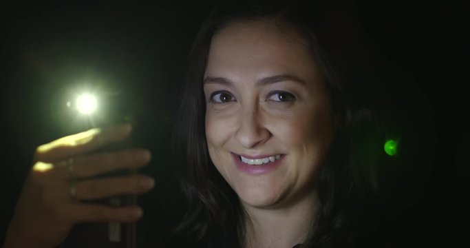 BCU young woman using cellphone at night, looks from screen to viewer and smiles. Hand-held 4K with lens flares, stabilized in camera.