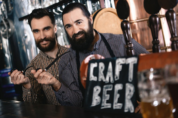 Two bearded men hold in hands tobacco smoking pipes, standing near sign with inscription Craft Beer.