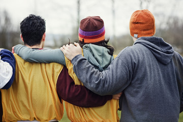 View from behind of three friends with their arms around one another in sports kit and woolly hats.