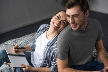 Happy man and woman in glasses writing in notepad while lying in bed