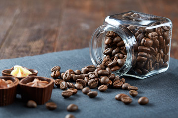 Side view of overturned glass jar with coffee beans and chocolate candies on wooden background, selective focus