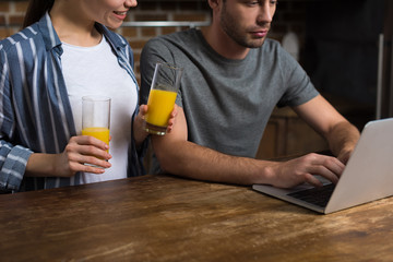 Young woman holding glasses with juice while man working on laptop