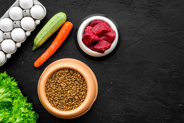 Dry pet food with natural ingredients. Raw meat, vegetables zucchini and carrot near eggs on black backgroud top view copy space