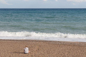 Fototapeta na wymiar Warm clear sunny summer day. A girl in a gray sweatshirt and jeans shorts sits with her back to the camera on the sad shore and looks at the sea