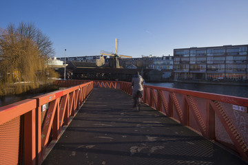 Obraz premium street life scene in Amsterdam West with warm sunrise light, with a cyclist in motion biking on a red bridge, with modern residential building and a historical old wooden windmill in the background