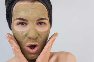 Fototapeta premium Closeup studio portrait of happy amazed young woman applying facial cosmetic clay organic mask on her face, wears black towel on hair. Female taking care of her face skin, isolated over white wall