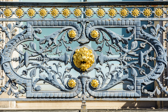 Close-up view of the wrought iron railing of the window of a parisian building decorated with a golden lion head in the middle surrounded by vegetal patterns and golden flowers.