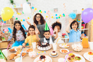 Group of children with color balloons sit behind festive table on which stands cake.