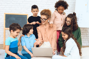 Nice teacher sits with school children who have surrounded her and are looking at laptop.