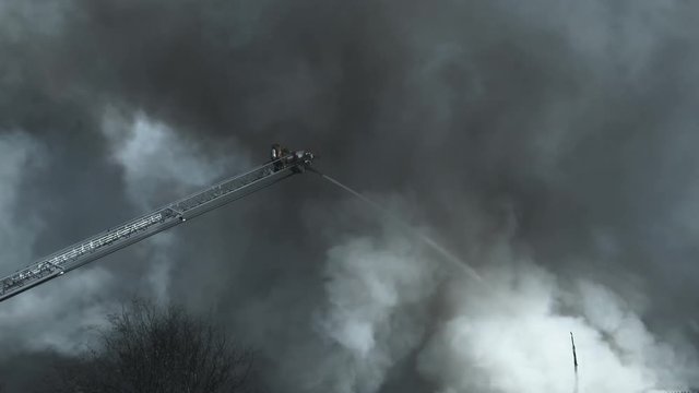 Zoom out to wide shot of fireman at the end of ladder using firehose to fight blaze below.